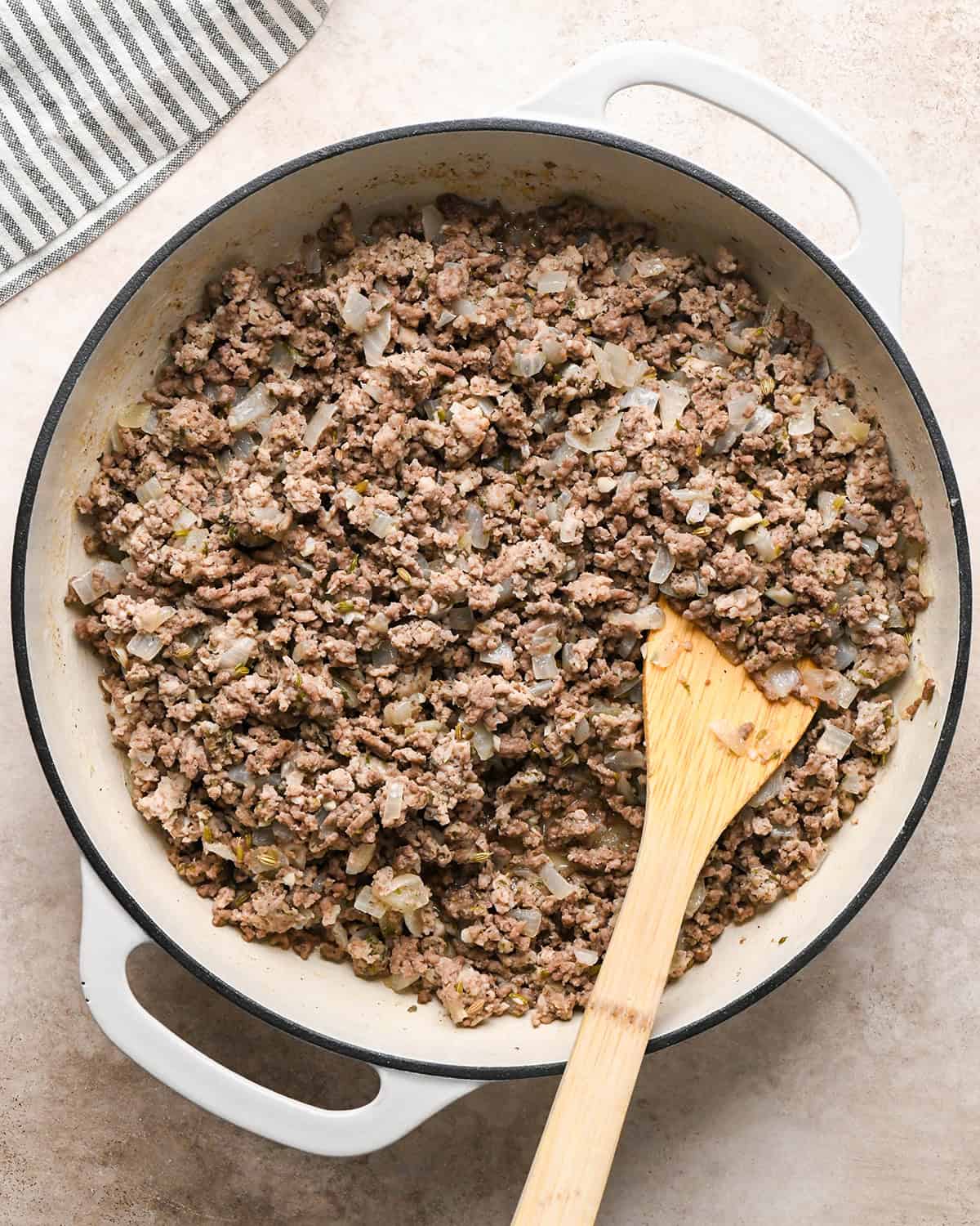Making baked ziti - onion, garlic, Italian sausage, and ground beef in a white saucepan, cooked and being stirred with a wooden spoon