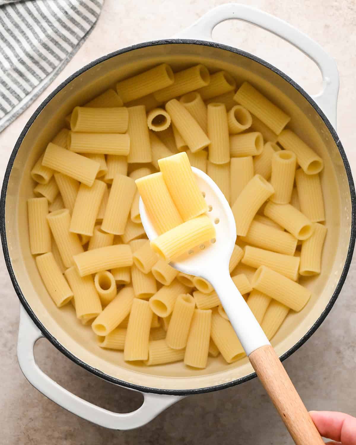 Cooked ziti pasta in a white saucepan, a serving spoon is picking up 3 noodles