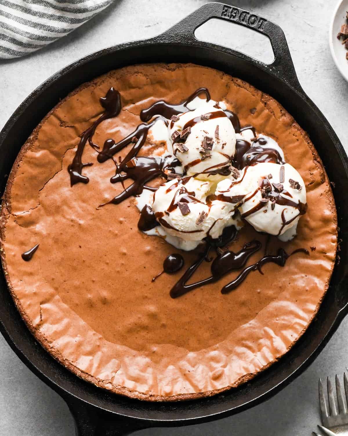 Making skillet brownies - overhead photo of brownies baked in a cast iron skillet and sitting on a countertop, topped with vanilla ice cream, chocolate shavings, and chocolate sauce