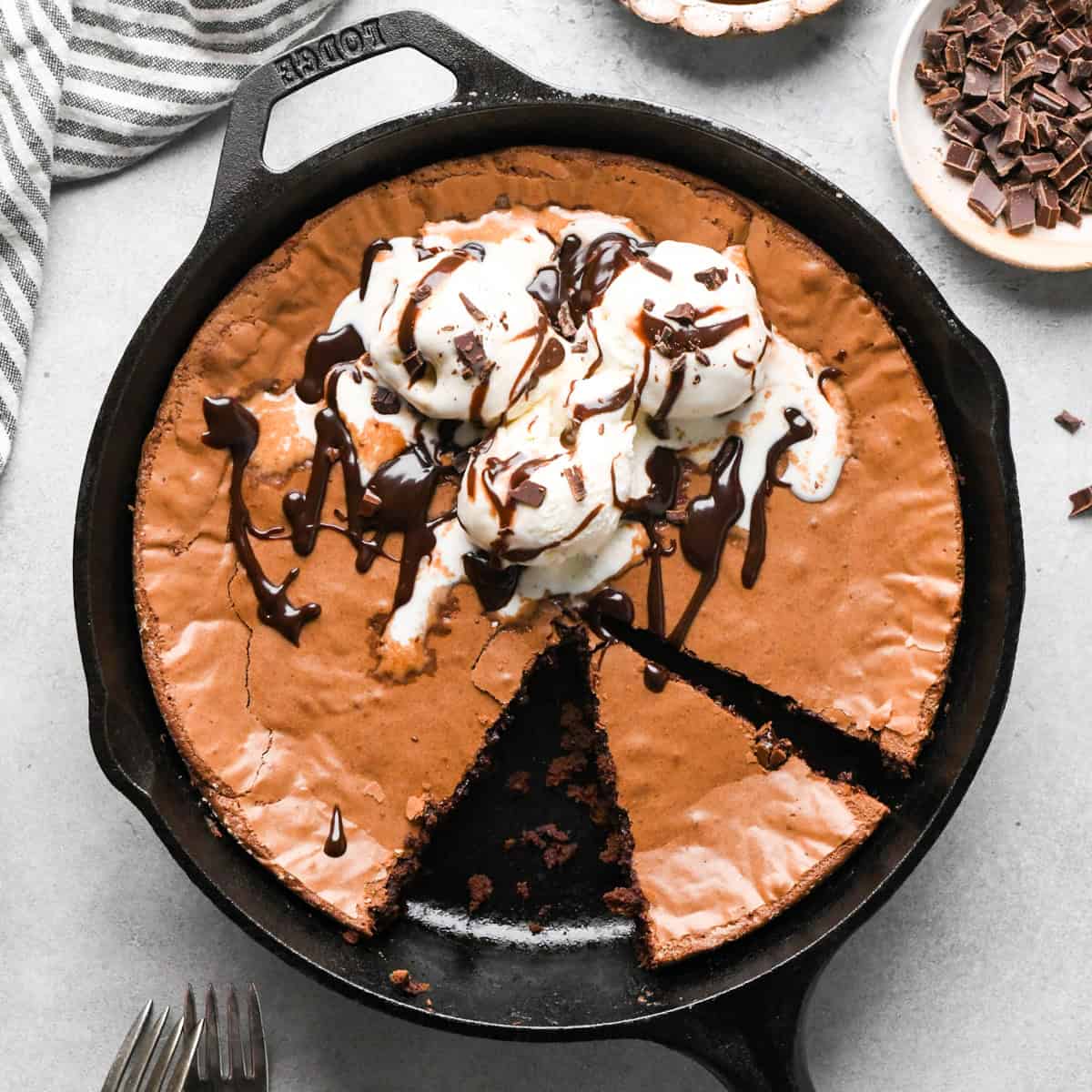 Cast iron skillet brownie topped with vanilla ice cream, chocolate sauce, and chocolate shavings, with a piece cut out of it, surrounded by a bowl of chopped chocolate and a striped towel. 