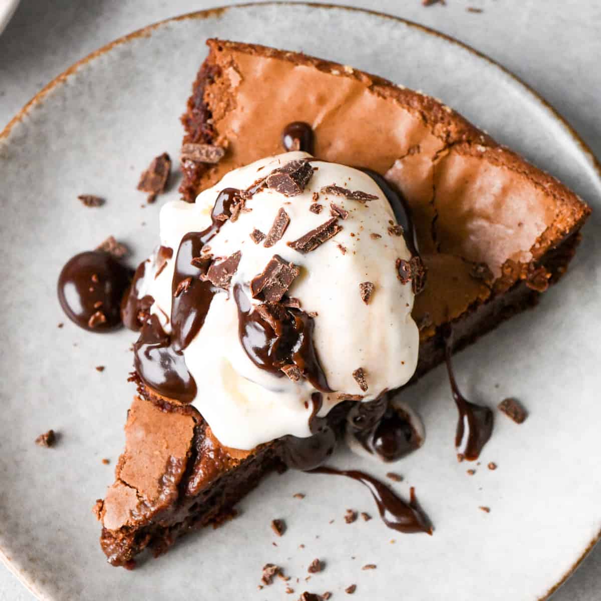 One slice of skillet brownie on a white/gray plate topped with vanilla ice cream, chocolate sauce, and chocolate shavings