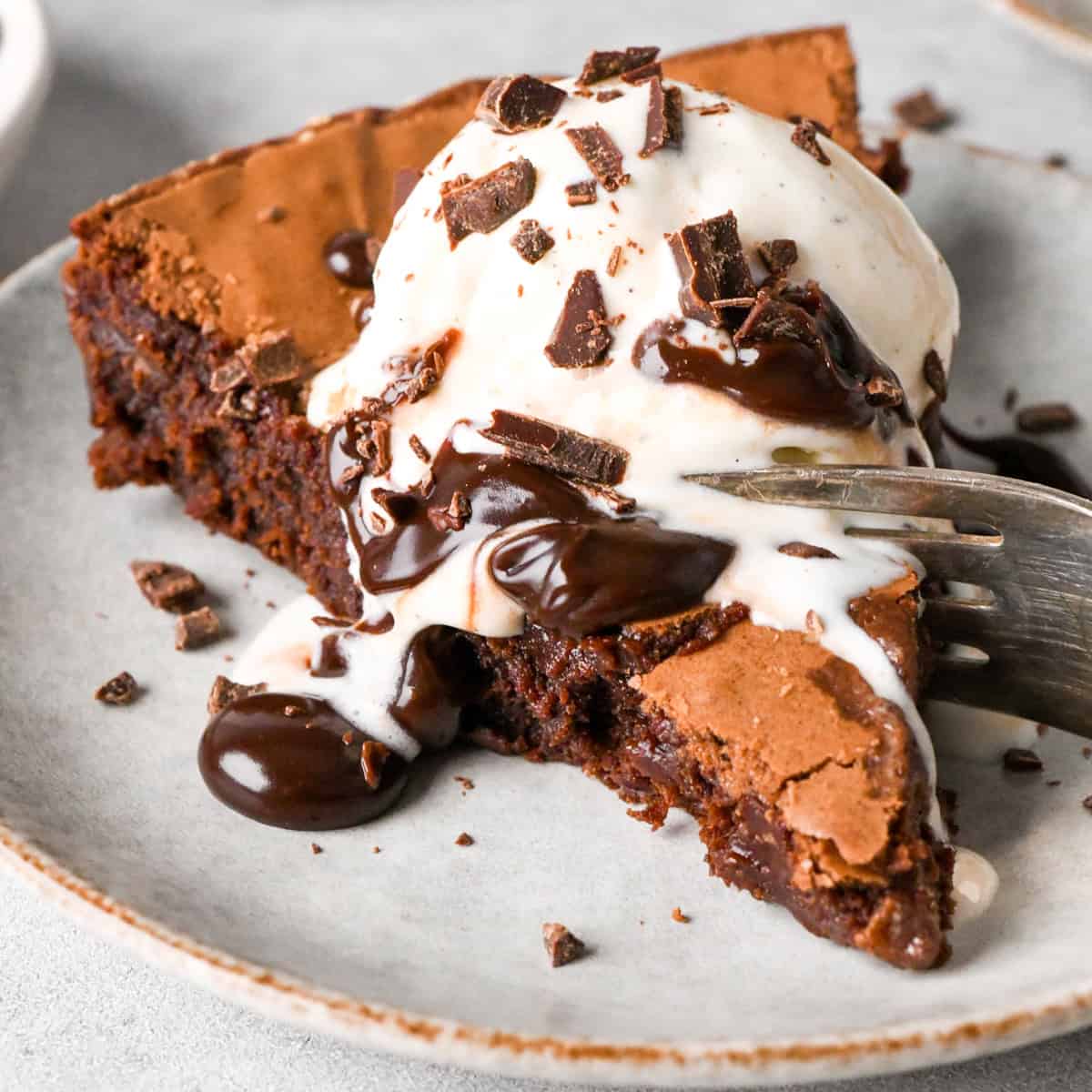 Close up photo of one slice of a cast iron skillet brownie topped with vanilla ice cream, chocolate sauce, and chocolate shavings and served on a white plate with a fork taking a bite out of the slice of brownie