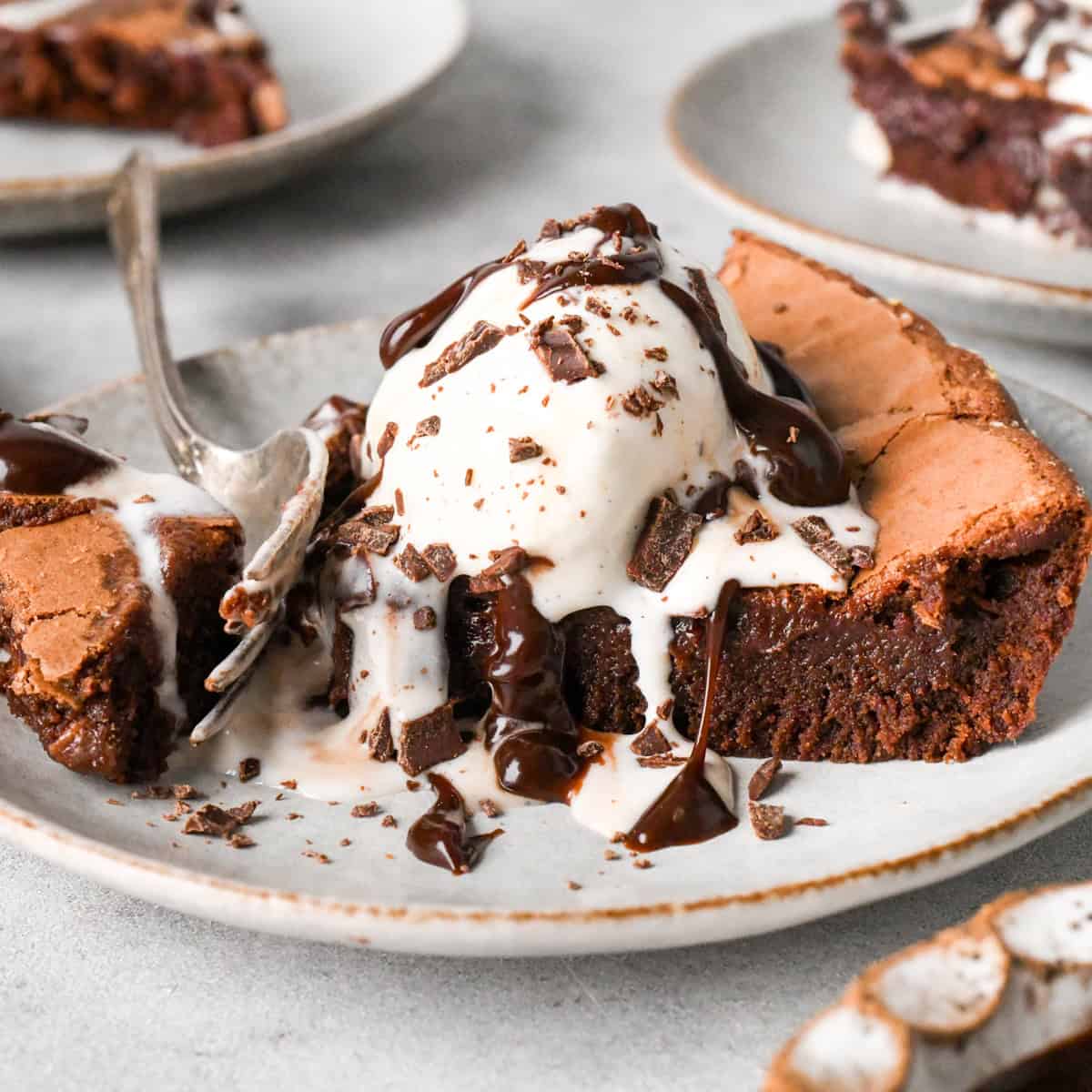 One slice of skillet brownie on a white/gray plate topped with vanilla ice cream, chocolate sauce, and chocolate shavings with one bite broken off by a fork