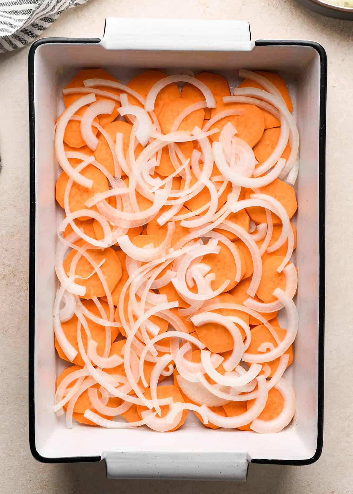 assembling sweet potato gratin - a layer of sliced onion put over a layer of sliced sweet potatoes in the bottom of a white baking dish with a black rim