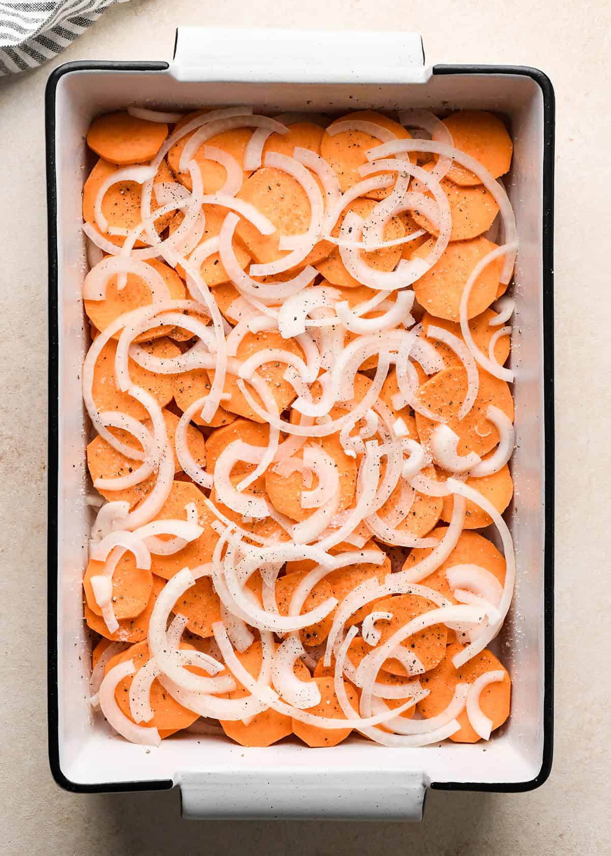 assembling sweet potato gratin - a layer of sliced onion put over a layer of sliced sweet potatoes in the bottom of a white baking dish with a black rim