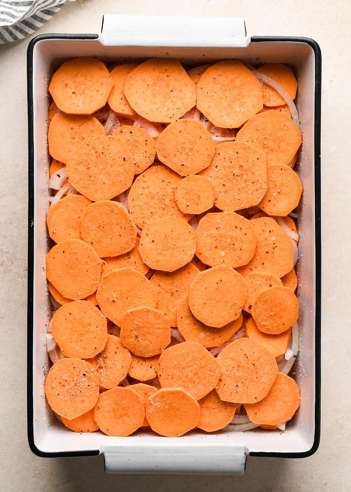 assembling sweet potato gratin - a layer of sliced sweet potatoes put over a layer of sliced onion in the bottom of a white baking dish with a black rim sprinkled with salt and pepper