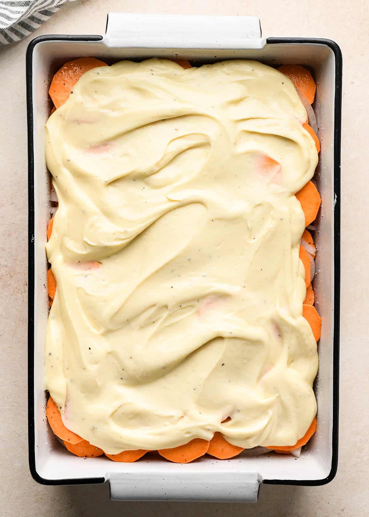 assembling sweet potato gratin - cheese sauce added to the layers of sweet potatoes and onion in a white baking dish with a black rim