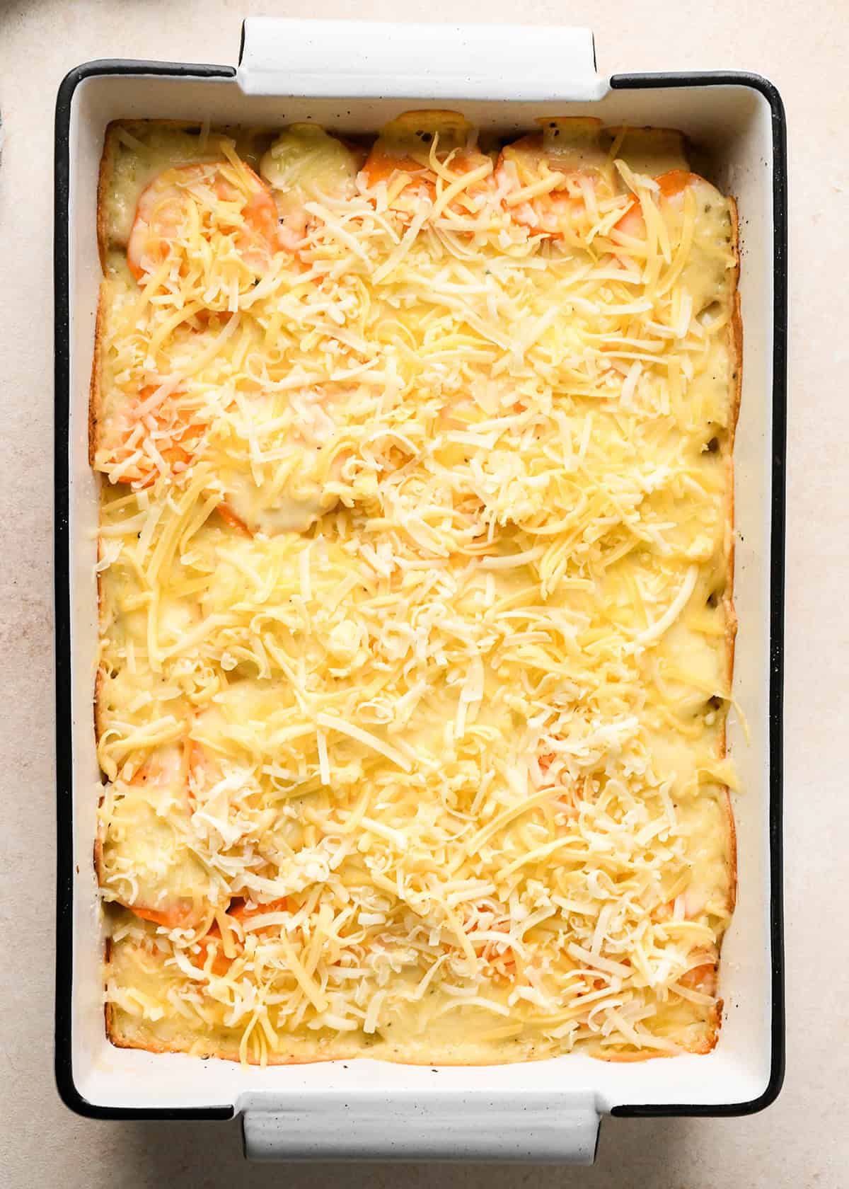 cheese sprinkled over the top of partially baked sweet potato gratin in a white baking dish with a black rim