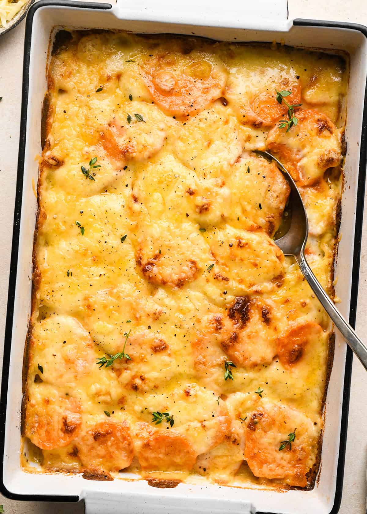 a silver spoon taking a scoop of sweet potato gratin in a baking dish after baking  topped with melted cheese, fresh thyme and black pepper