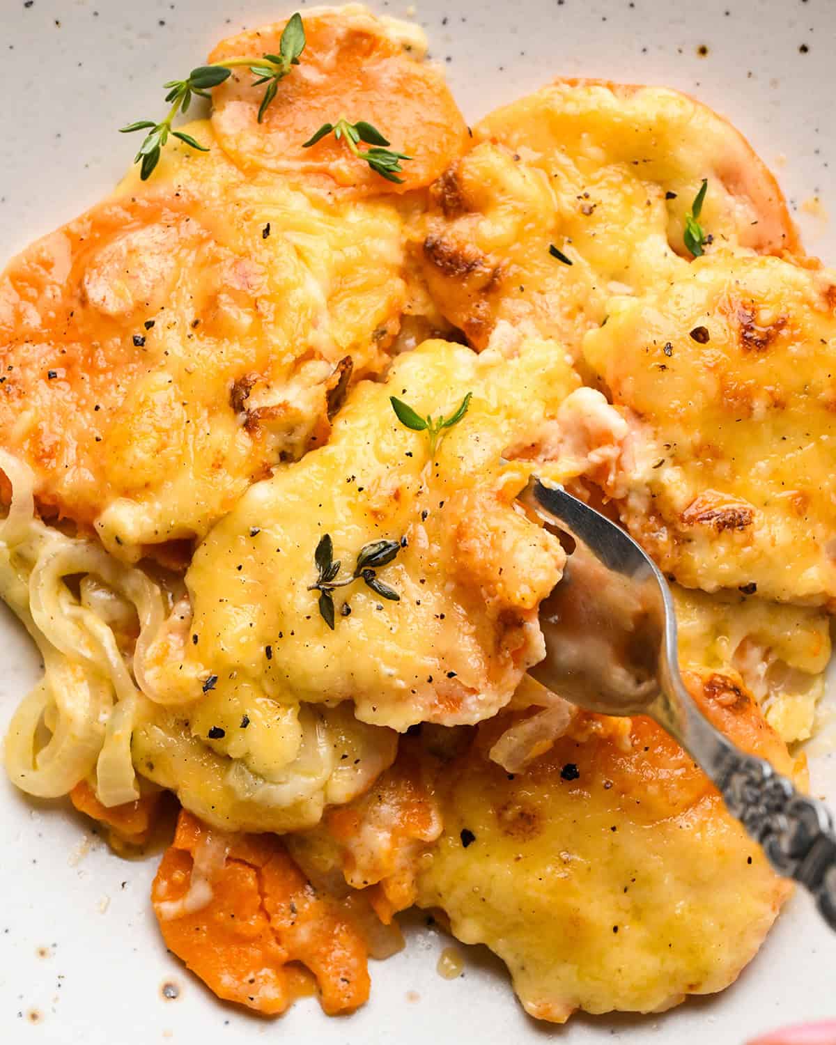 a fork taking a bite of a serving of sweet potato gratin on a plate topped with melted cheese, fresh thyme and black pepper