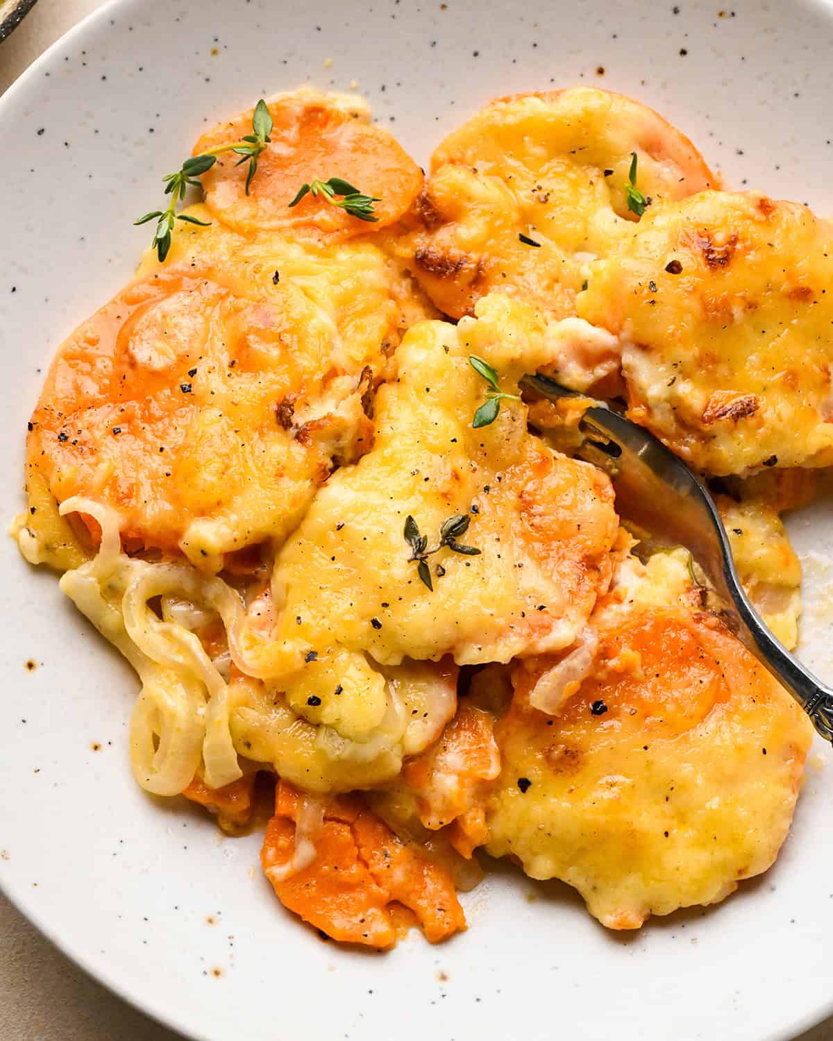 a fork taking a bite of a serving of sweet potato gratin on a plate topped with melted cheese, fresh thyme and black pepper
