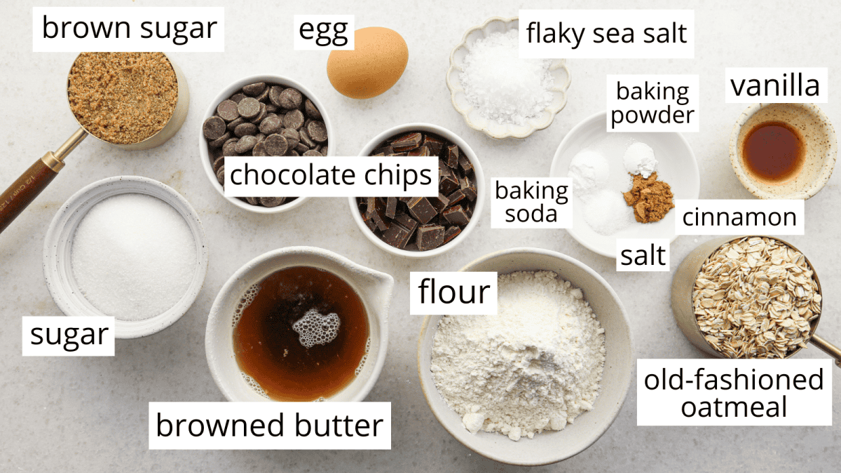 Top-down view of measured ingredients for brown butter oatmeal cookies, including old-fashioned oats, flour, browned butter, chocolate chips, brown sugar, granulated sugar, an egg, vanilla extract, baking soda, baking powder, cinnamon, salt, and flaky sea salt.
