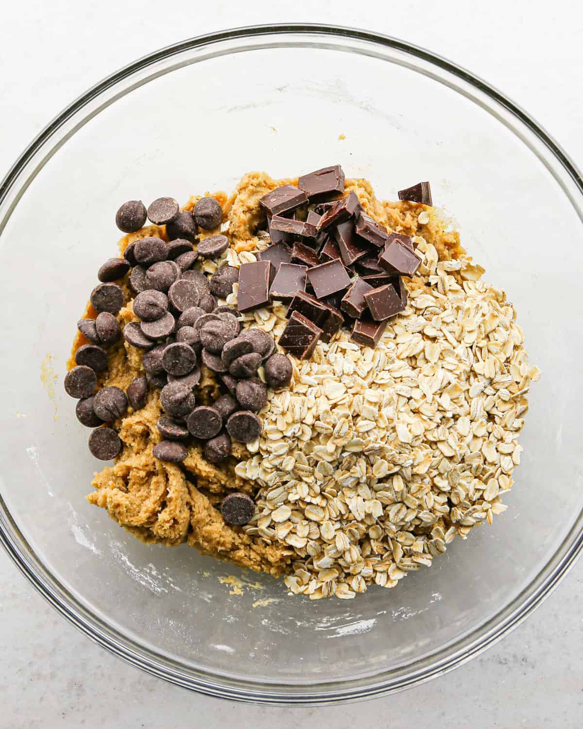 Making brown butter oatmeal cookies - overhead photo of oats, chocolate chips, and chocolate chunks added to the cookie dough in a clear bowl.