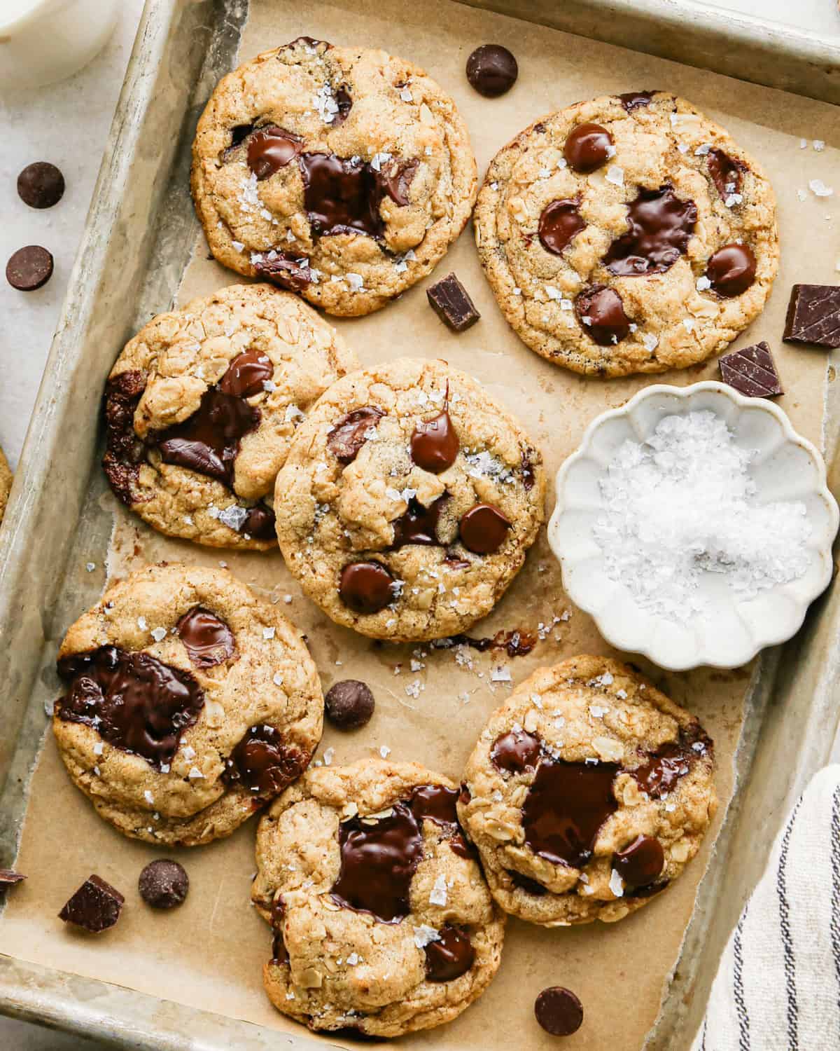 Overhead photo of a completed batch of brown butter oatmeal cookies on a baking sheet with parchment paper, surrounded by additional chocolate chunks and a little white bowl of flaky sea salt.