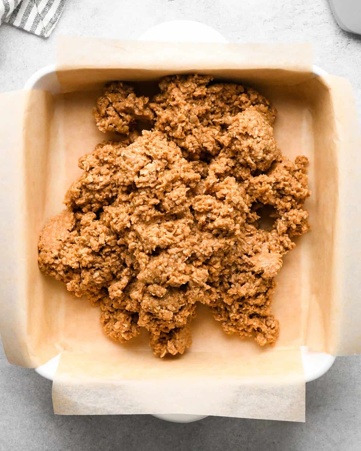 Making peanut butter oatmeal bars - overhead photo of peanut butter oatmeal bar mixture being poured into a white casserole dish lined with parchment paper.