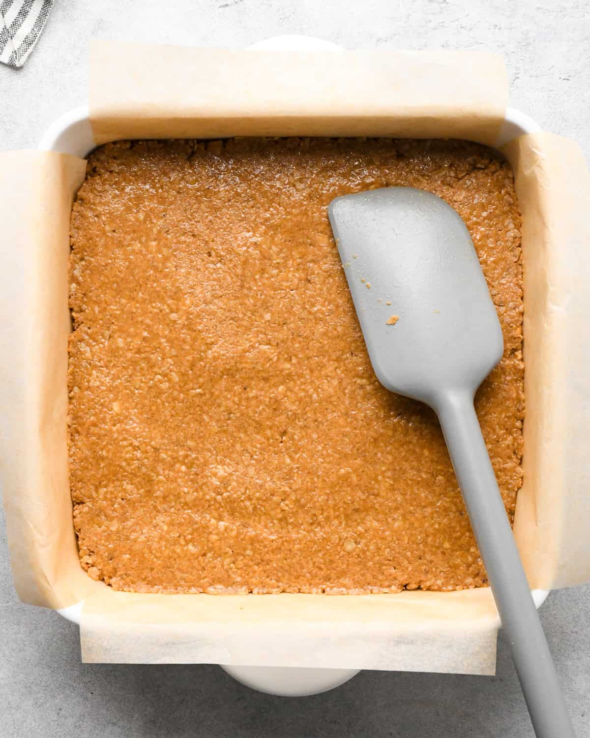 Making peanut butter oatmeal bars - overhead photo of peanut butter oatmeal bar mixture in a white casserole dish lined with parchment paper, being spread out with a gray spatula.