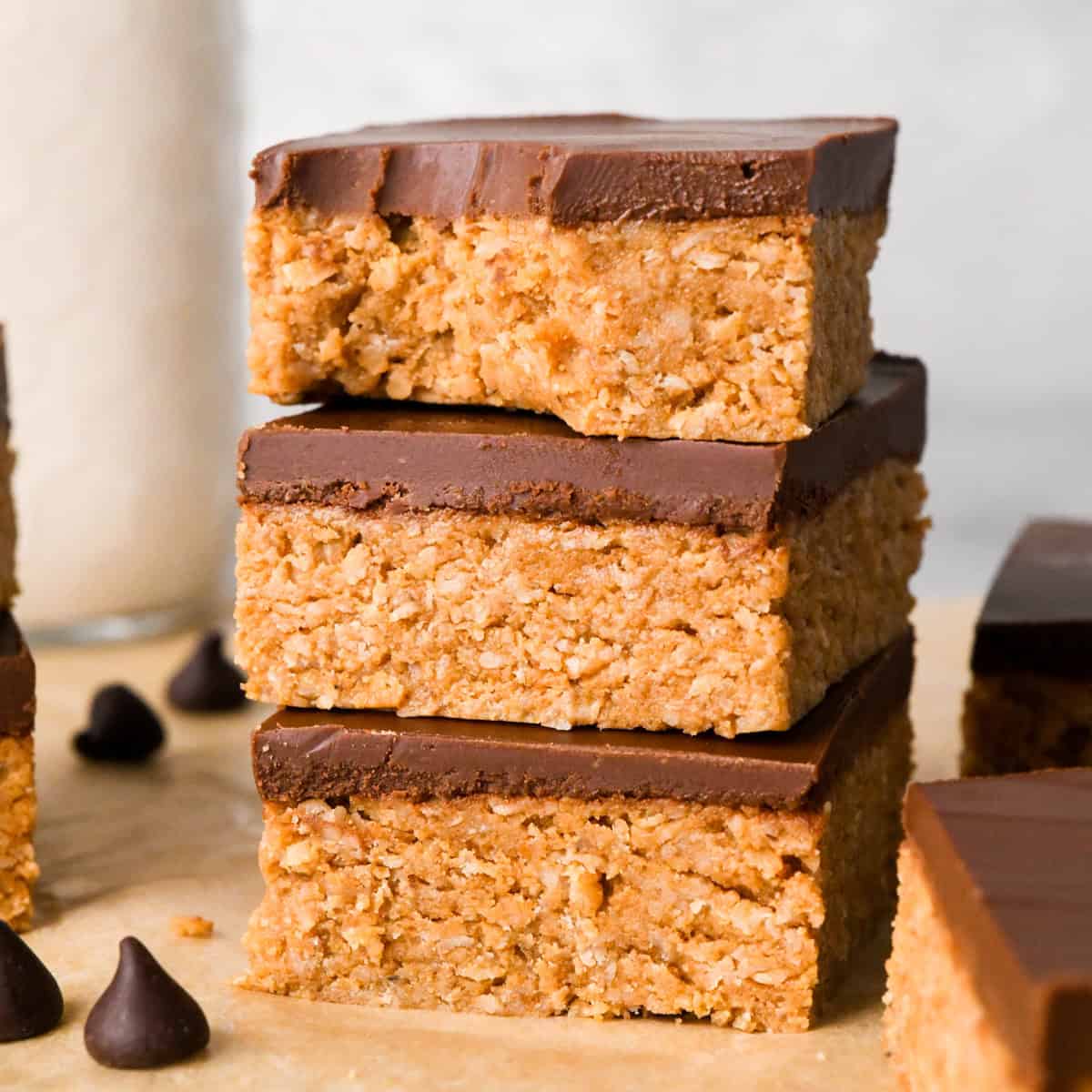 Close-up image of 3 peanut butter oatmeal bars topped with a smooth layer of chocolate and sitting on parchment paper, a bite is taken out of the top bar.