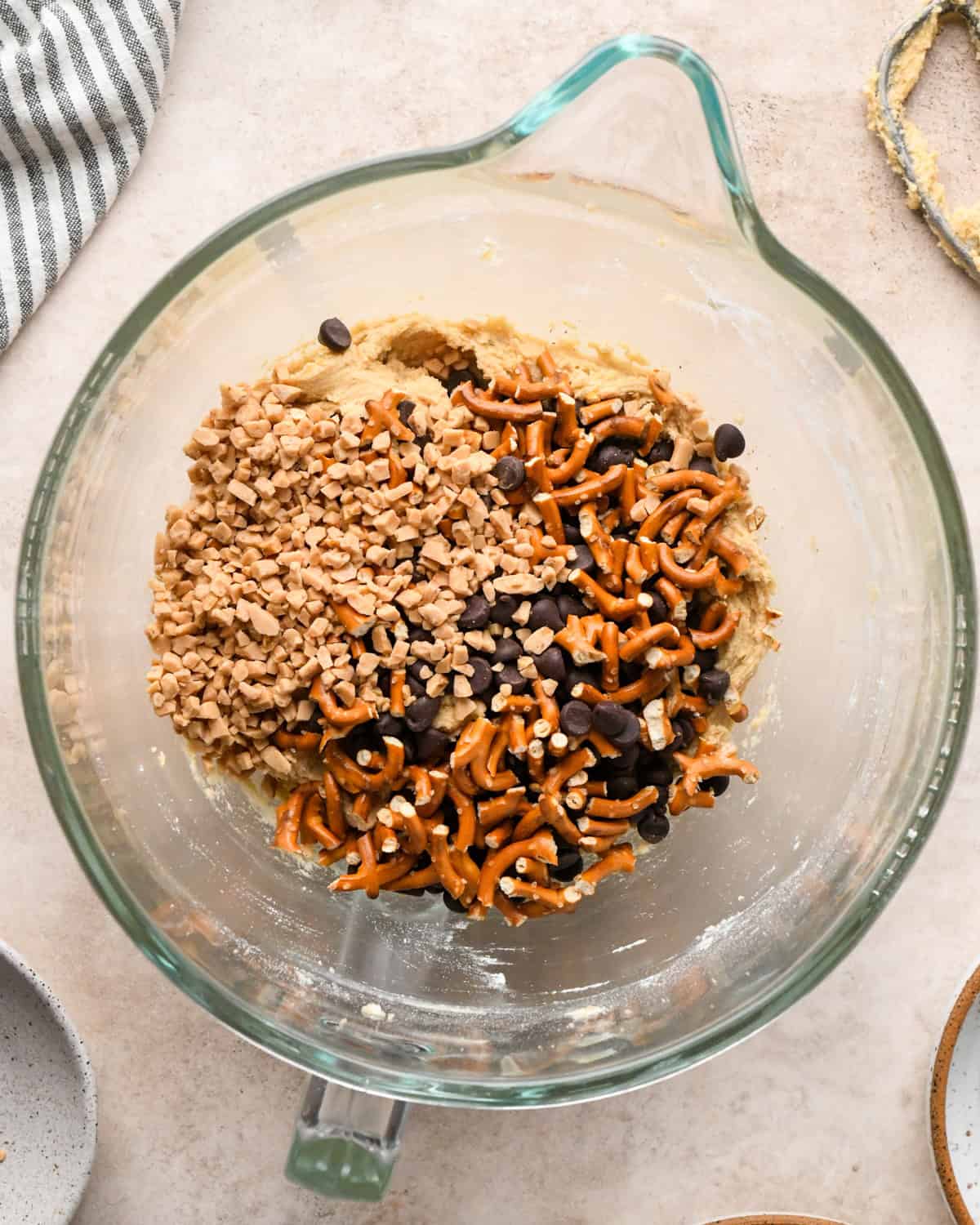 Making kitchen sink cookies - overhead photo of pretzel pieces, toffee pieces, and chocolate chips being added to cookie dough in a clear bowl.