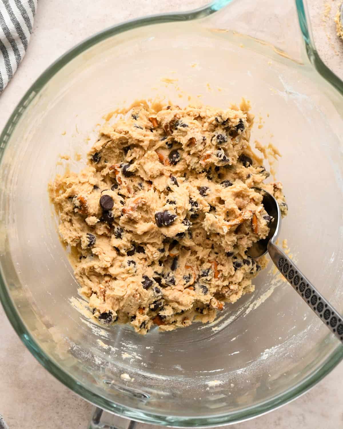 Making kitchen sink cookies - overhead photo of pretzel pieces, toffee pieces, and chocolate chips being added to cookie dough in a clear bowl and mixed together. A silver spoon is in the bowl.