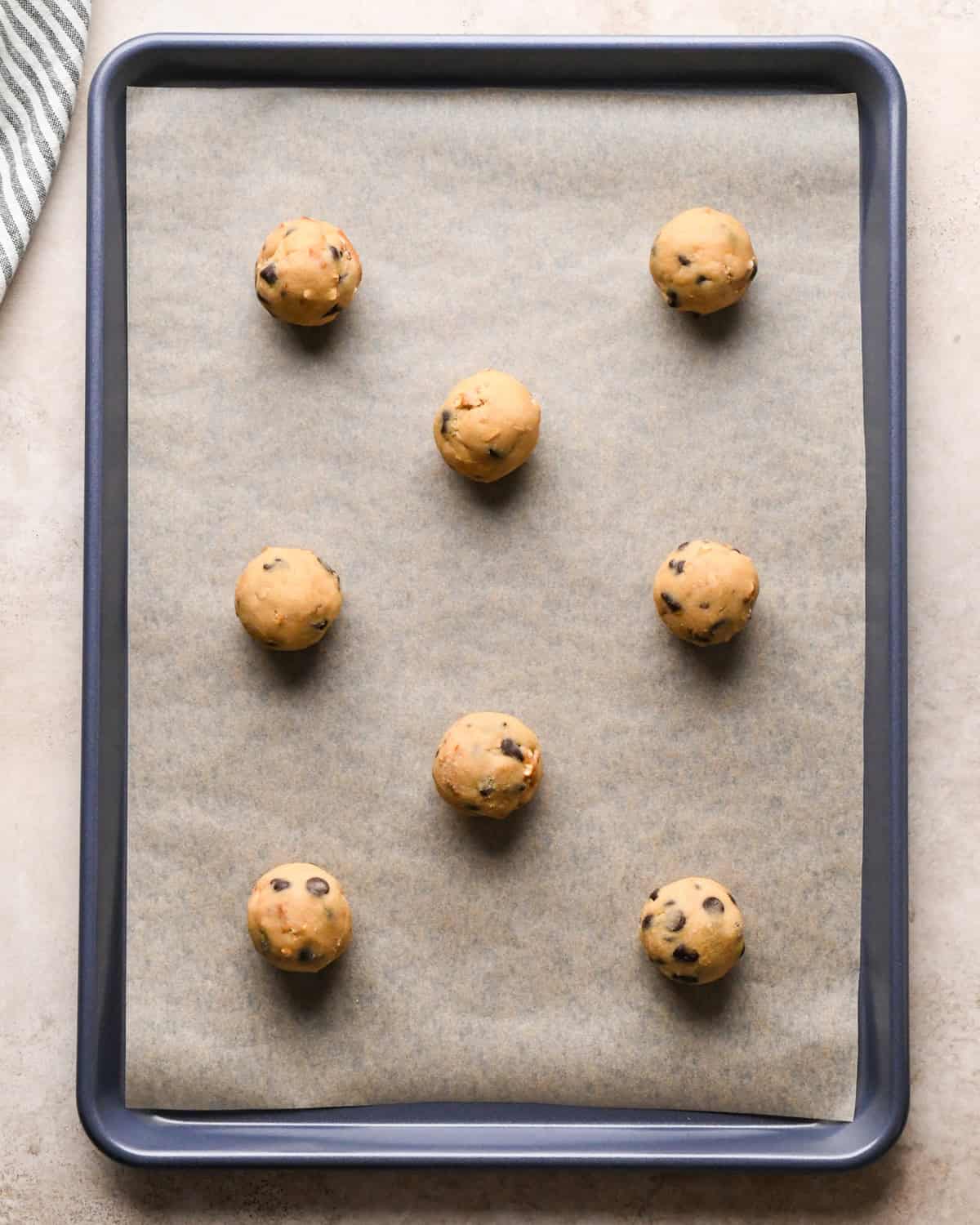 Making kitchen sink cookies - overhead photo of 8 rolled-up balls of cookie dough on a parchment-paper-covered baking sheet.