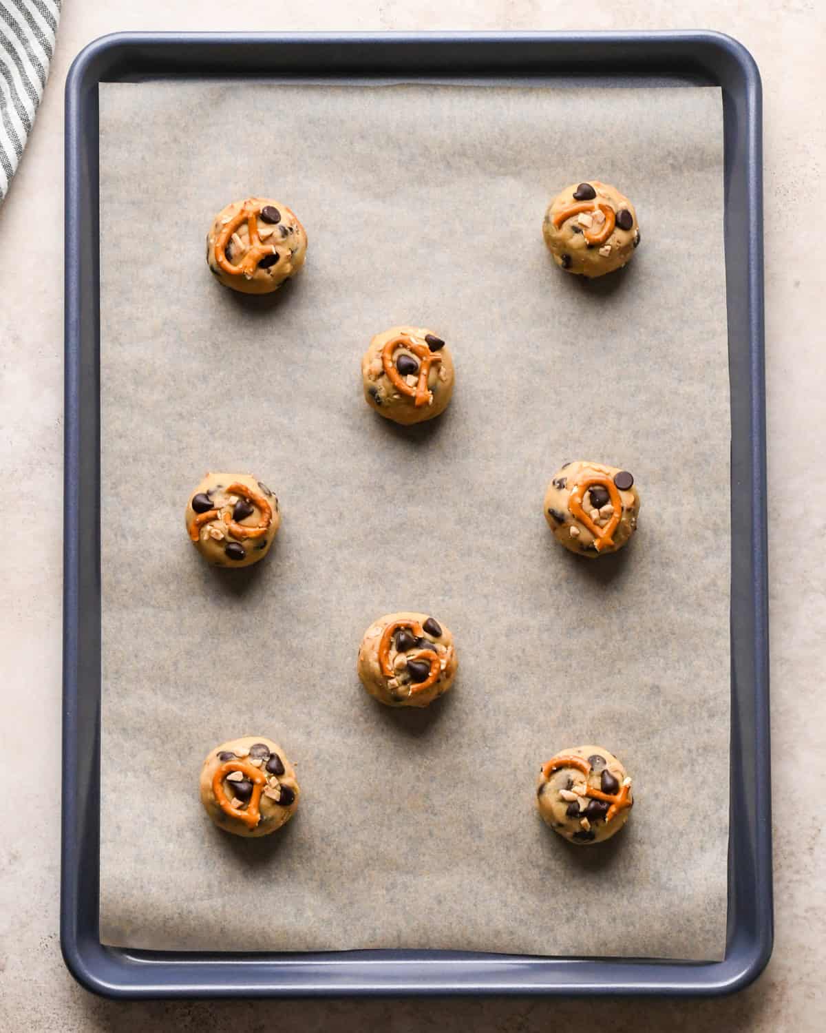 Making kitchen sink cookies - overhead photo of 8 rolled-up balls of cookie dough on a parchment-paper-covered baking sheet, with an additional pretzel piece placed on top of each cookie.