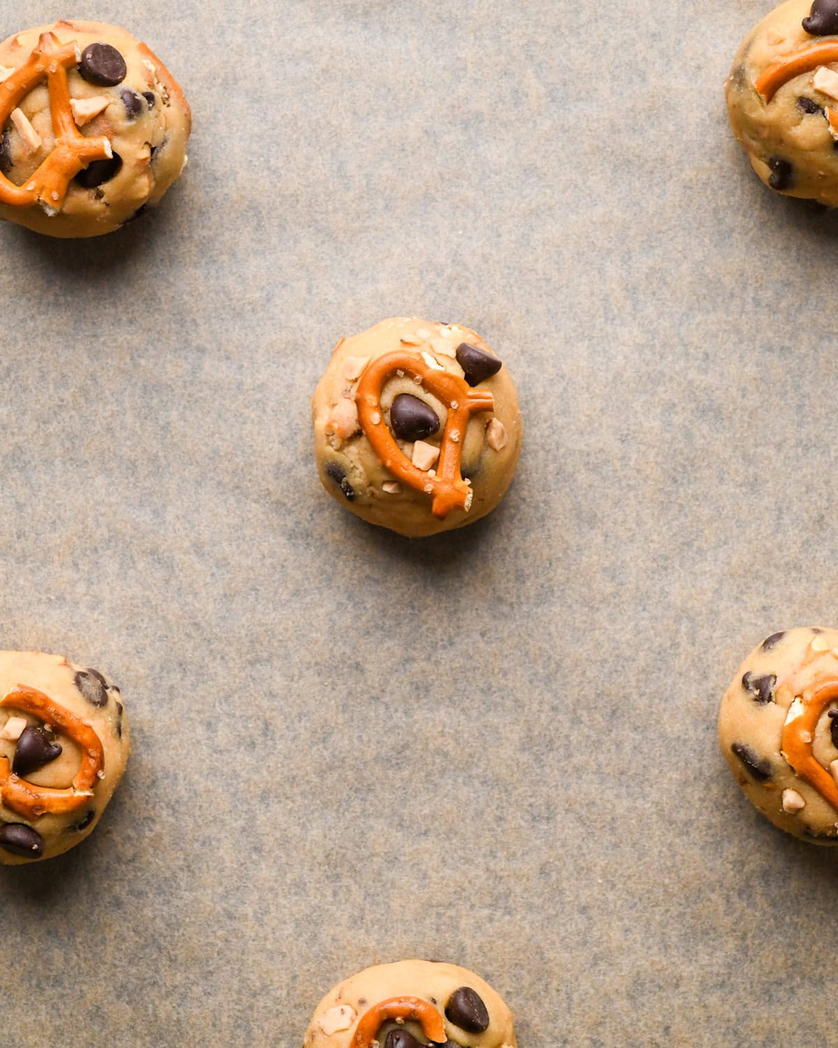 Making kitchen sink cookies - close-up photo of balls of cookie dough on a parchment-paper-covered baking sheet, with an additional pretzel piece placed on top of each cookie.