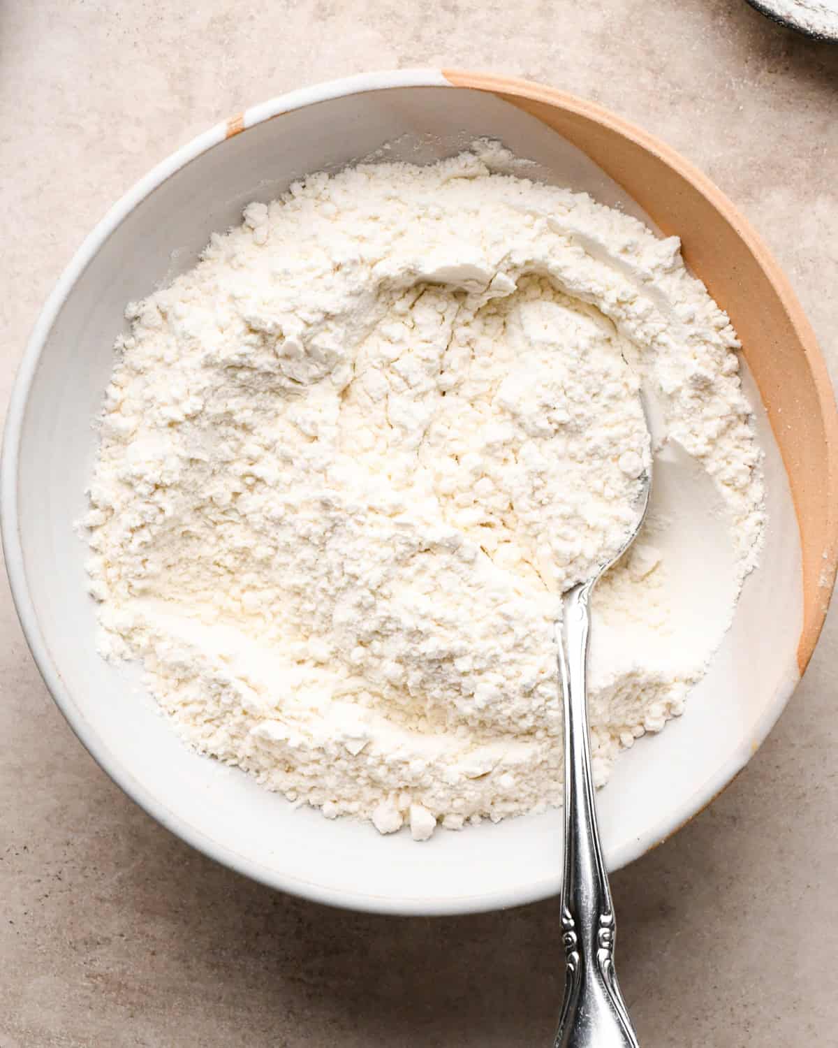 Making kitchen sink cookies - overhead photo of dry ingredients being added to a white-and-orange mixing bowl and stirred together with a silver spoon.