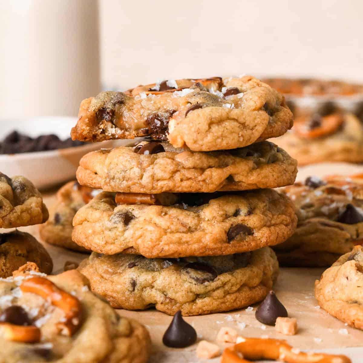 Close-up photo of a stack of 4 kitchen sink cookies loaded with chocolate chips, pretzel pieces, and bits of toffee, surrounded by additional cookies on parchment paper.