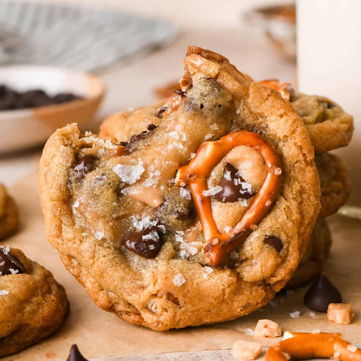 Close-up photo of kitchen sink cookies loaded with chocolate chips, pretzel pieces, and bits of toffee, topped with flaky sea salt. A bite is taken out of one cookie.