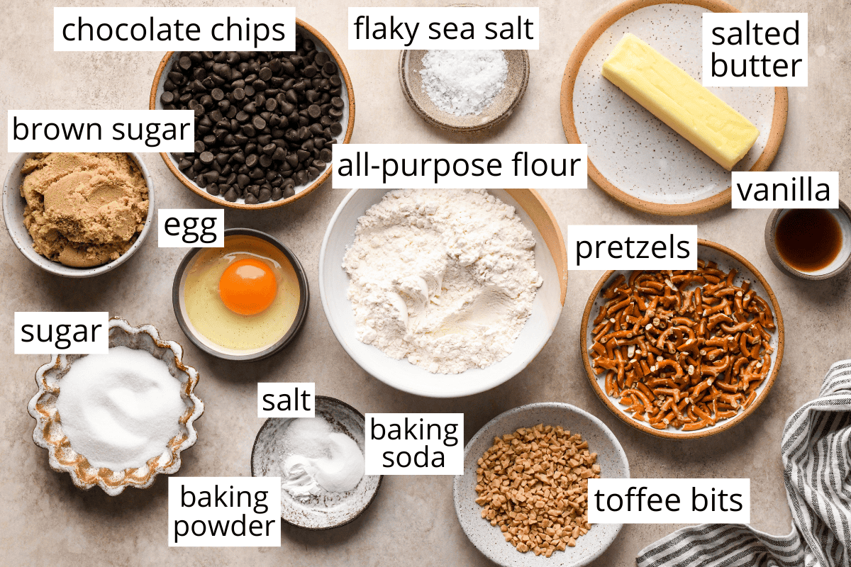 Top-down view of measured ingredients for kitchen sink cookies, including bowls of all-purpose flour, chocolate chips, pretzels, toffee bits, brown sugar, granulated sugar, salted butter, an egg, vanilla extract, baking soda, baking powder, salt, and flaky sea salt.