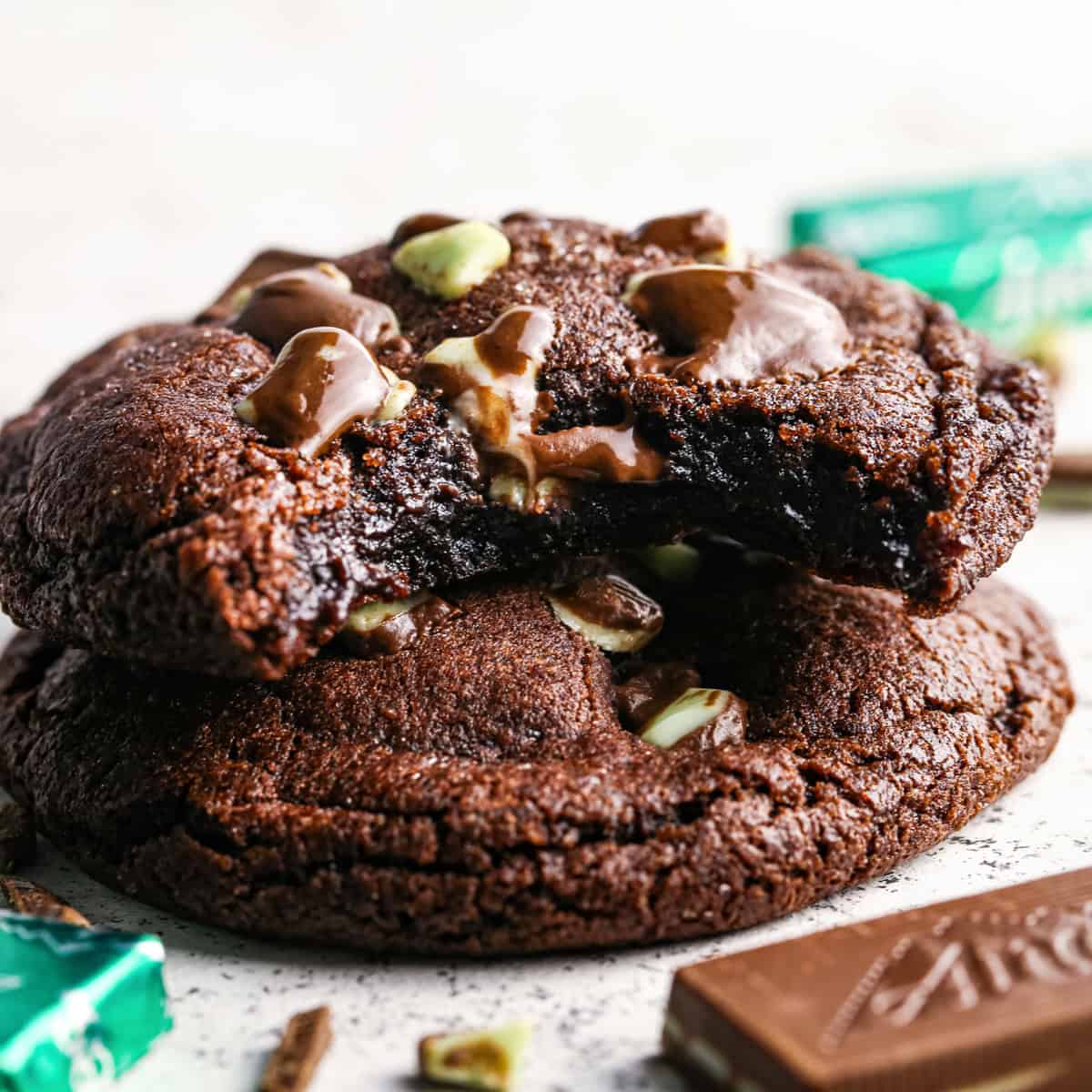 Close-up photo of a stack of 2 chocolate mint cookies with a bite taken out of the top cookie, surrounded by additional chopped mint chocolate.