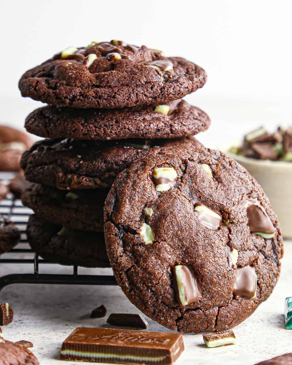 a stack of 5 chocolate mint cookies on a wire cooling rack with a sixth mint chocolate cookie leaning on the stack in front of the stack. Surrounded by more cookies and andes mints.