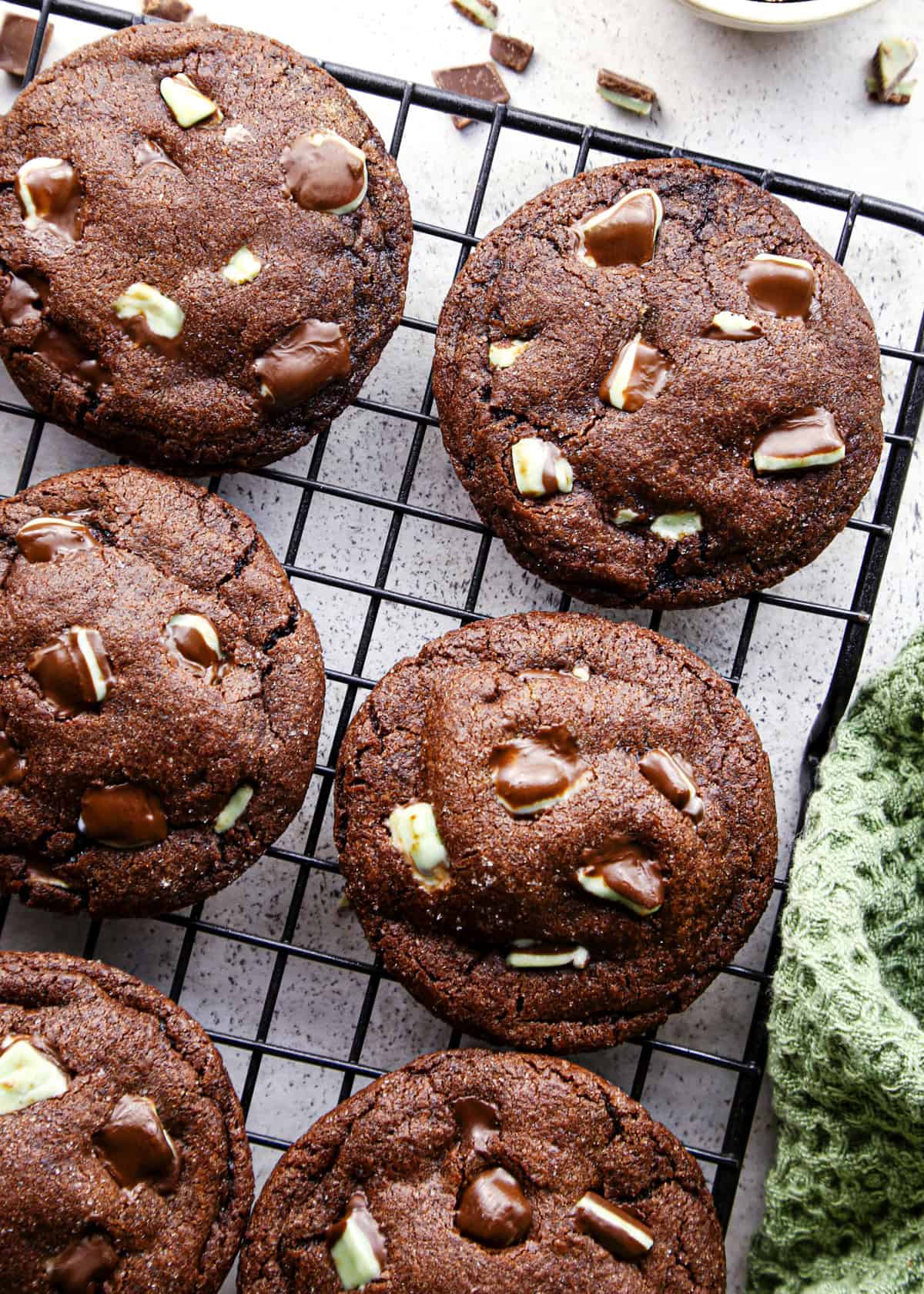 Overhead photo of chocolate mint cookies baked and cooling on a cooling rack, surrounded by additional mint chocolate pieces and a green towel on a countertop.