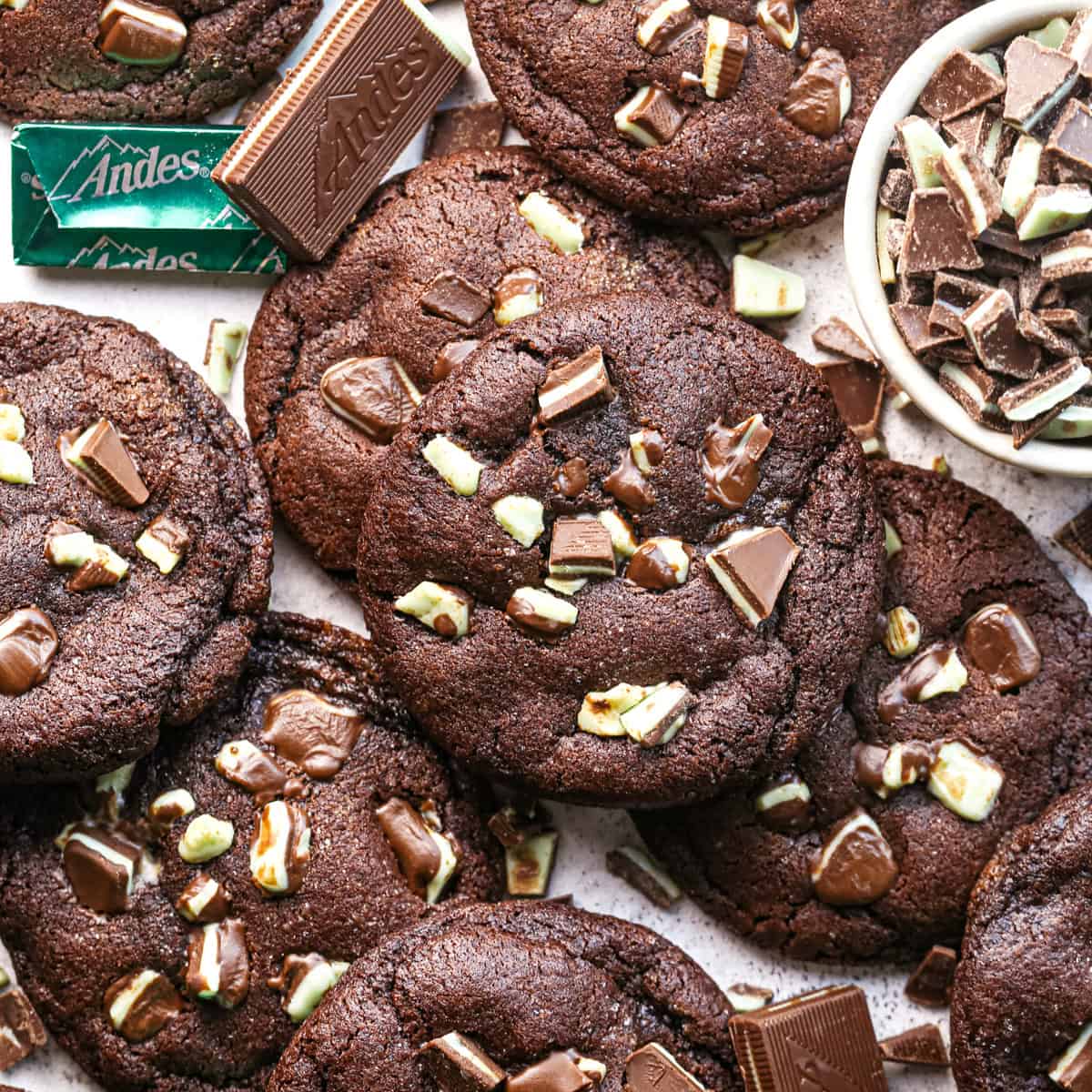 Close-up photo of rich chocolate mint cookies topped with chunks of chocolate and green mint candy pieces, surrounded by additional chopped mint chocolate.