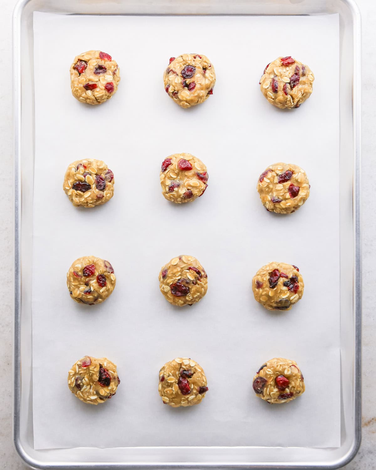 Making cranberry oatmeal cookies - overhead photo of 12 cranberry oatmeal cookie dough balls on a parchment-paper-covered baking sheet, all 12 cookies have been pressed down by hand.