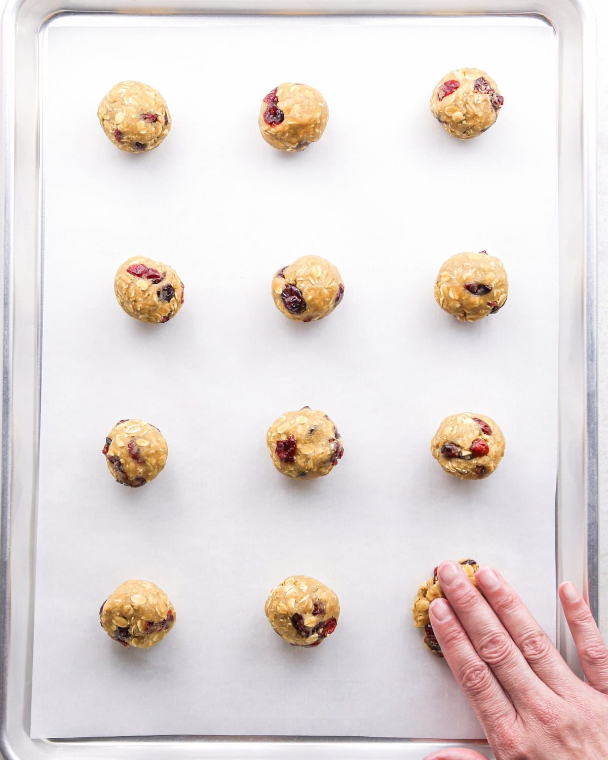 Making cranberry oatmeal cookies - overhead photo of 12 cranberry oatmeal cookie dough balls on a parchment-paper-covered baking sheet and being pressed down by hand.