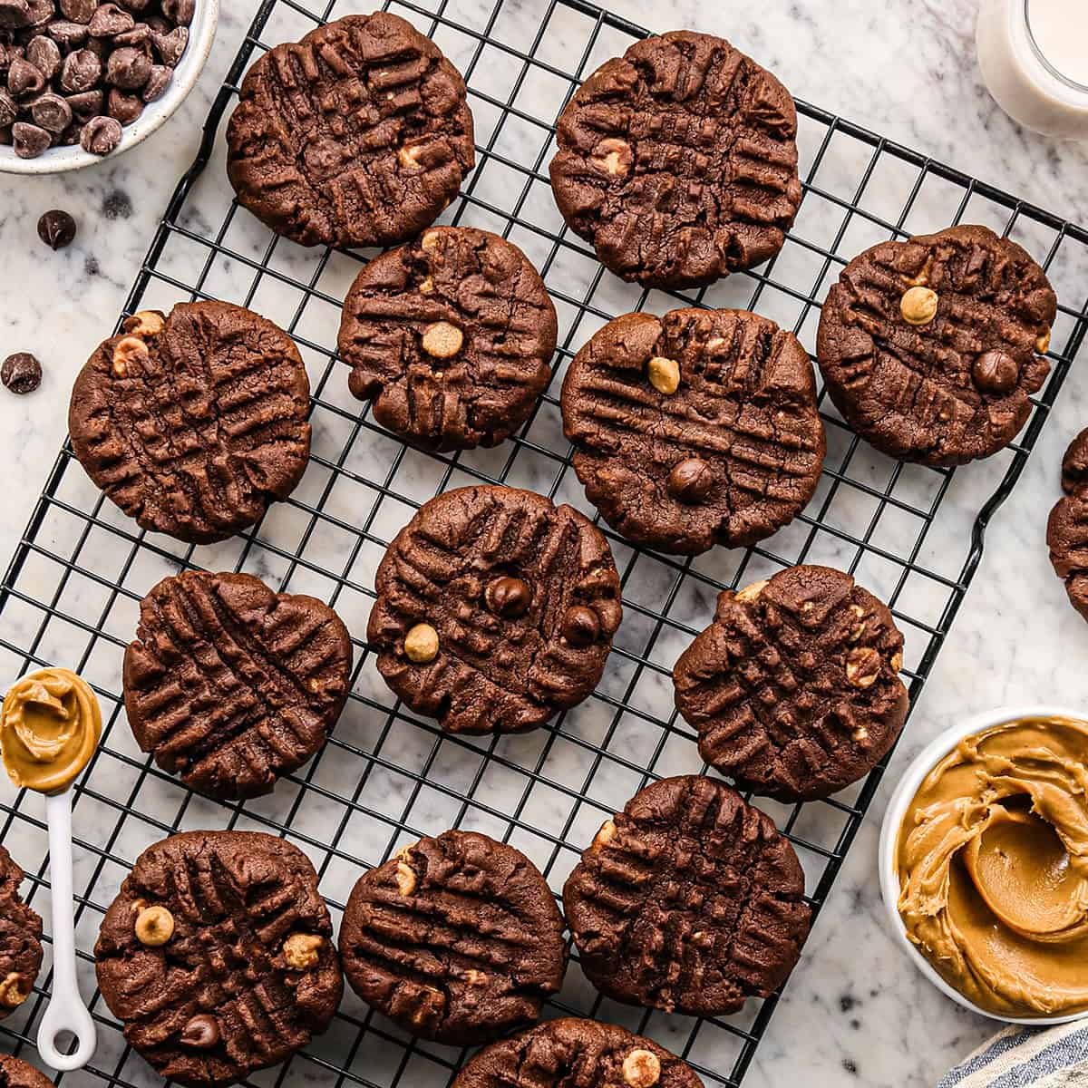 Overhead photo of chocolate peanut butter chip cookies on a cooling rack surrounded by bowls of chocolate chips and peanut butter.