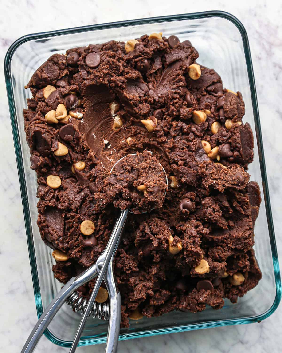 Overhead photo of chocolate peanut butter chip cookie dough in a container, chilled and being scooped with a cookie scoop. 