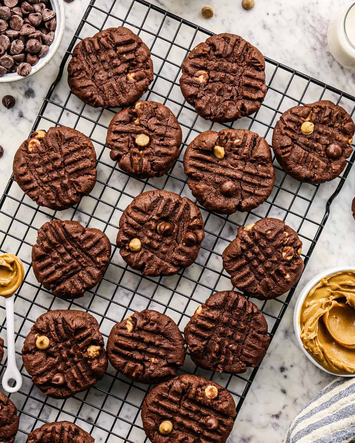 Overhead photo of chocolate peanut butter chip cookies on a cooling rack surrounded by bowls of chocolate chips and peanut butter.