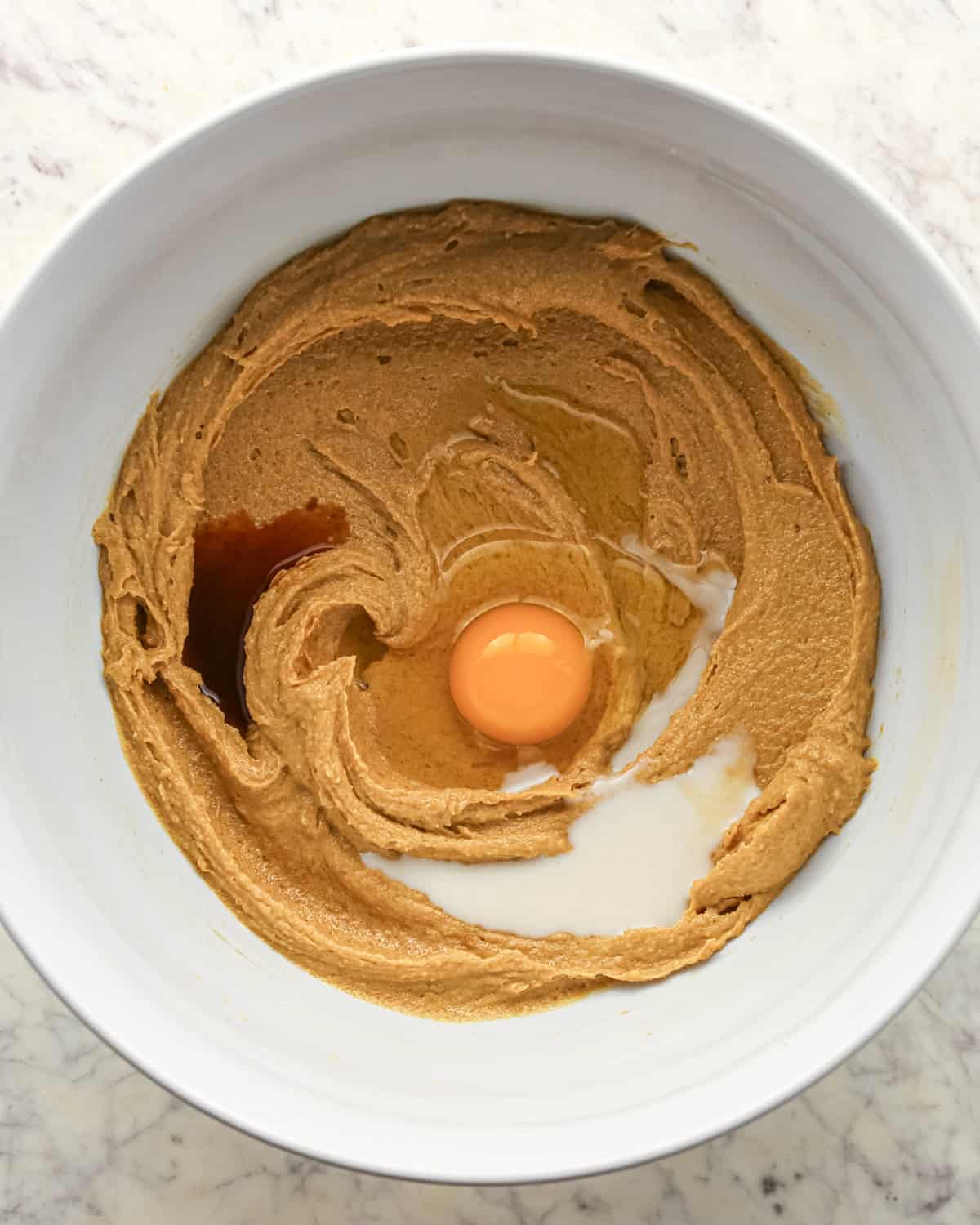 Making chocolate peanut butter chip cookies - vanilla, egg, and milk are added to a white bowl with butter, peanut butter, sugar, and brown sugar.