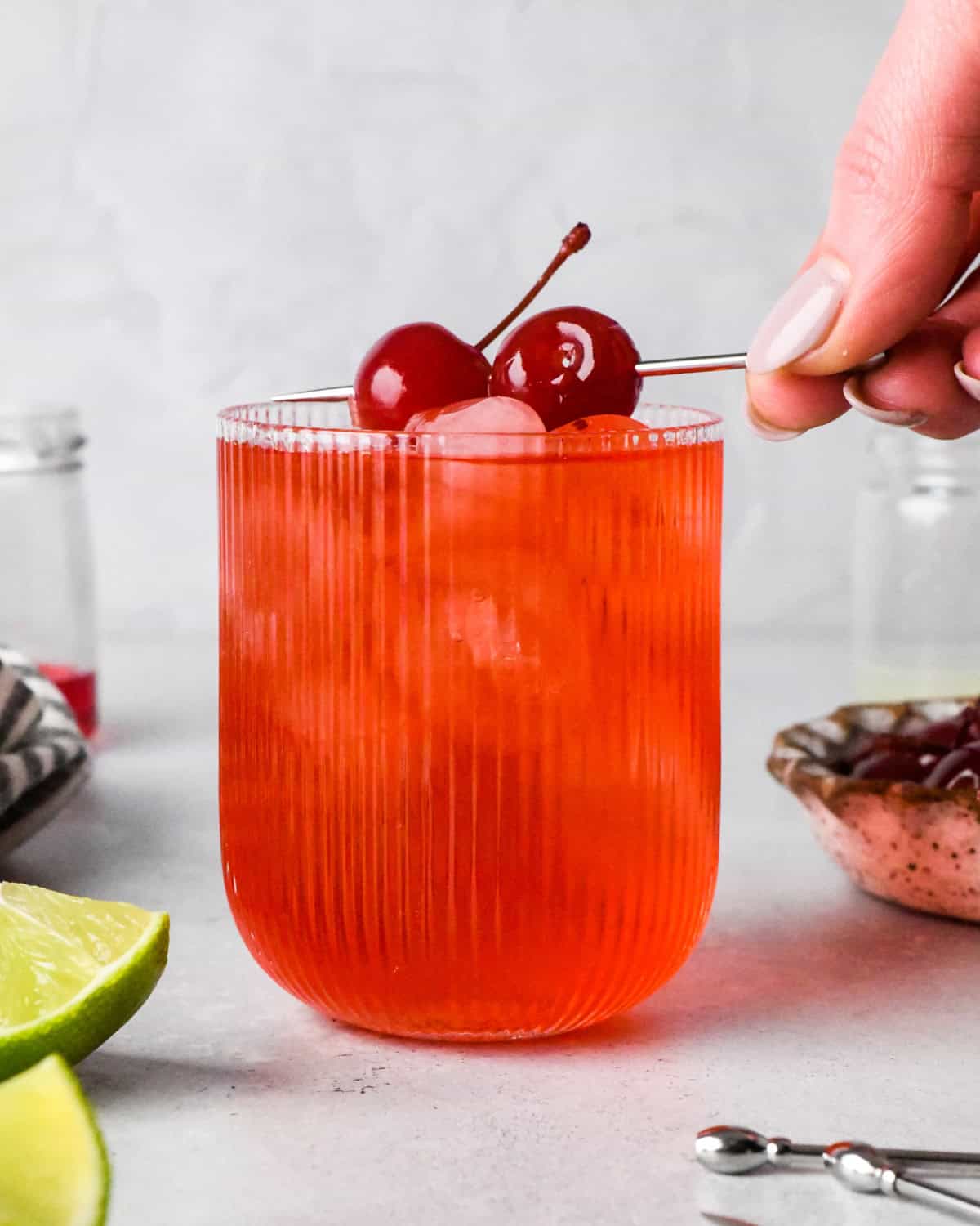 Making a Shirley Temple - close-up photo of grenadine, lime juice, and ginger ale in a clear glass with ice, and a person is placing 2 maraschino cherries on top.
