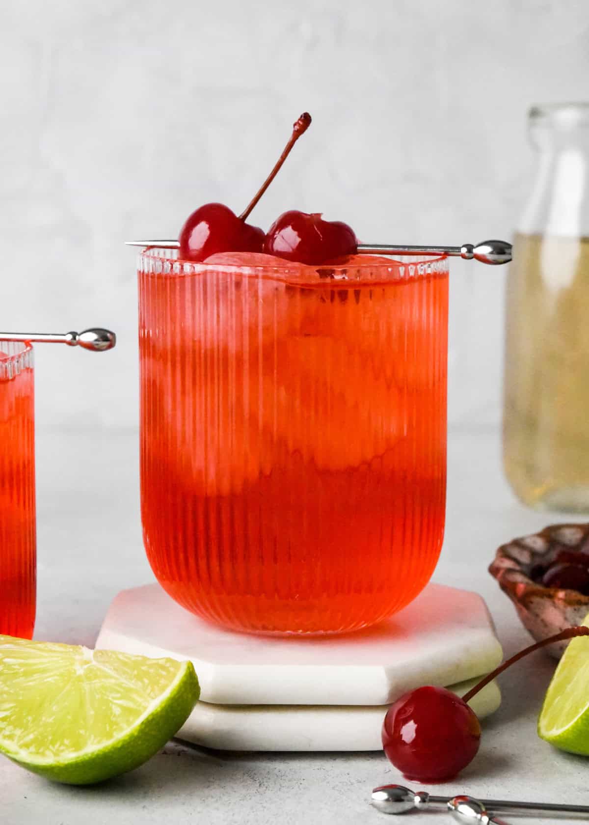 Close-up photo of a Shirley Temple served over ice in a glass, garnished with a maraschino cherry on a cocktail pick. The glass is sitting on a coaster and surrounded by lime wedges and maraschino cherries.