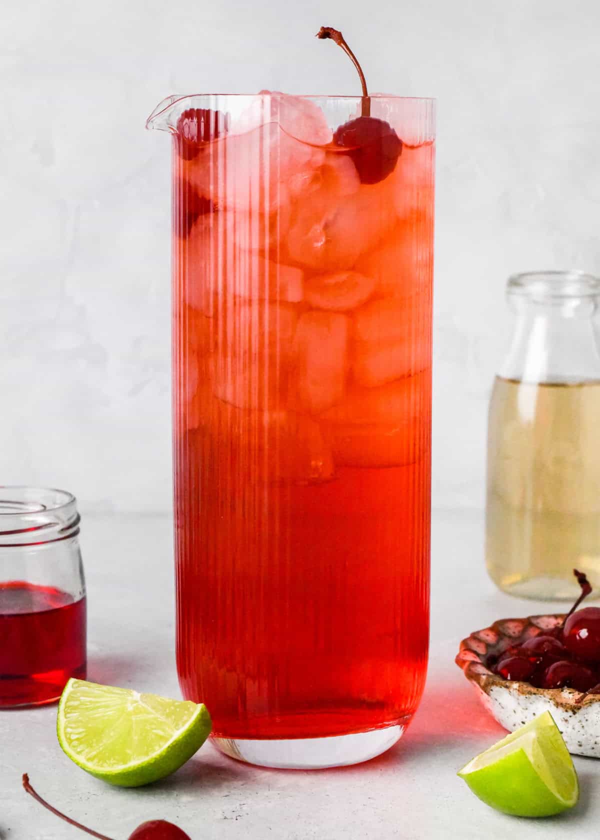 Making a large-batch Shirley Temple - close-up photo of grenadine, lime juice, and ginger ale in a large clear serving glass with ice and 2 maraschino cherries on top. The glass is sitting on a light countertop next to extra ingredients.