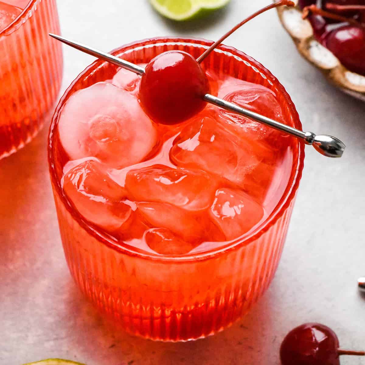 Close-up photo of a Shirley Temple served over ice in a glass, garnished with a maraschino cherry on a cocktail pick.