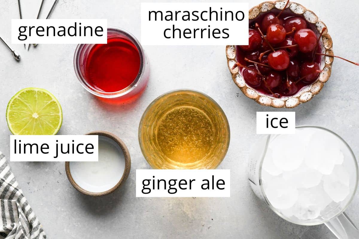 Top-down view of ingredients for a Shirley Temple, including grenadine, maraschino cherries, ice, lime juice, ginger ale, and a halved lime on a light surface.