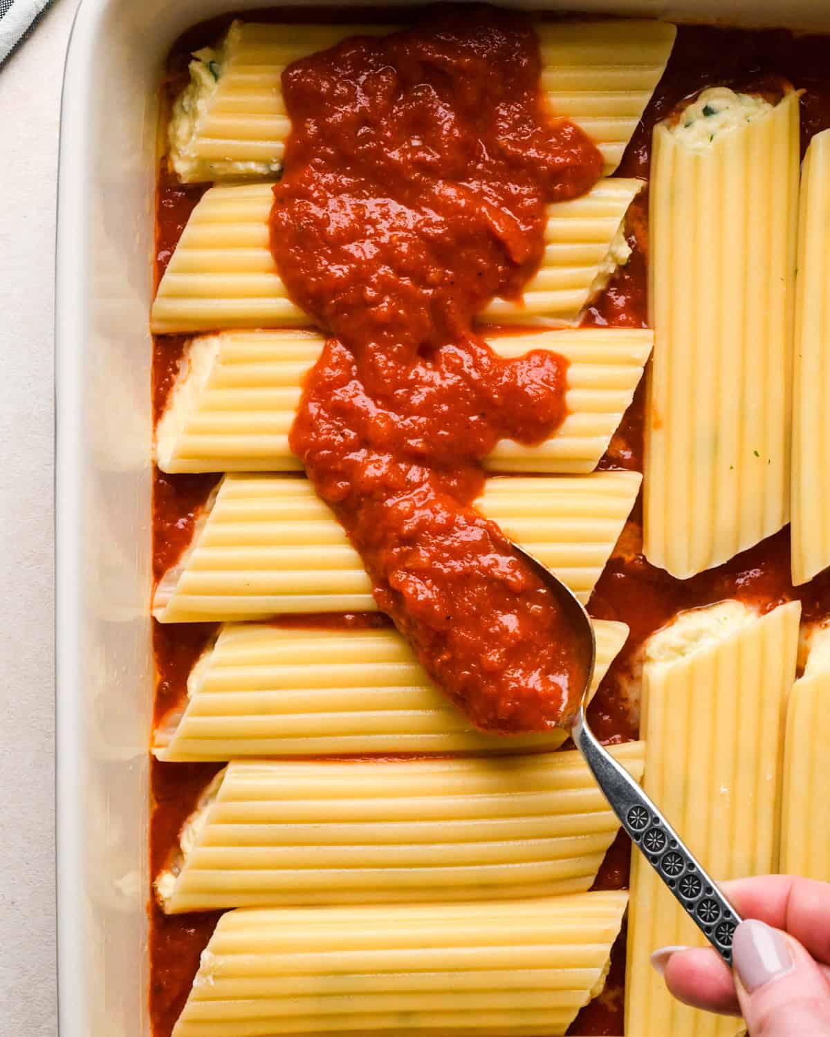 Making stuffed manicotti - overhead photo of a person pouring marinara sauce over stuffed manicotti noodles in a casserole dish.