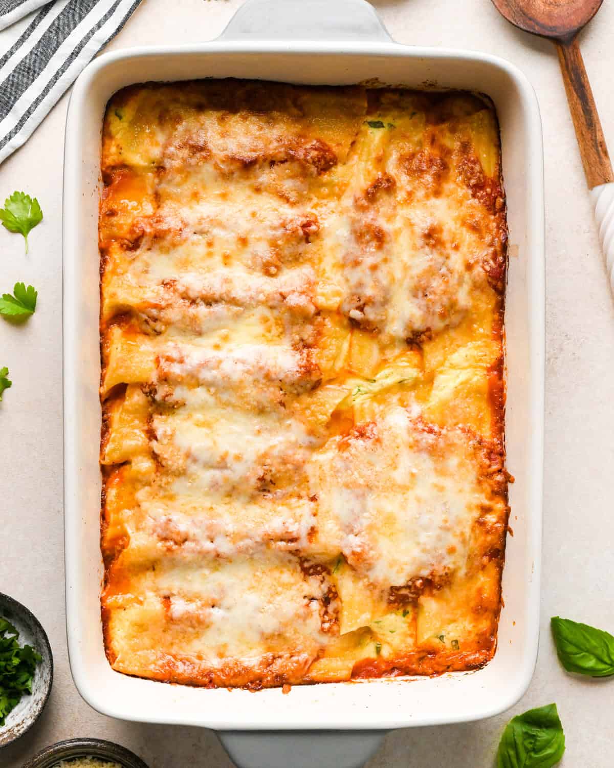 Overhead view of stuffed manicotti baked in a white casserole dish. The casserole dish is shown next to fresh basil leaves and a wooden serving spoon.