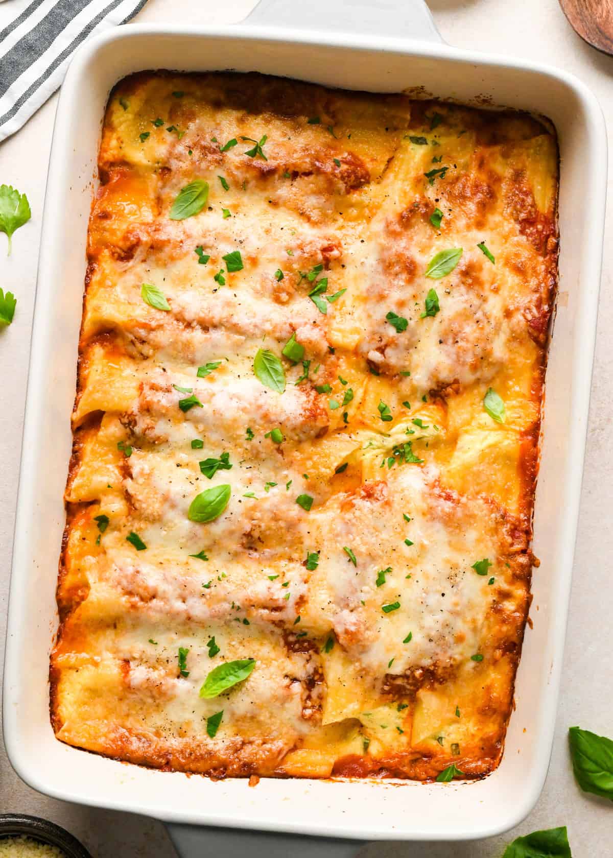 Overhead view of stuffed manicotti baked in a white casserole dish. The entree is shown with golden brown cheese and topped with fresh basil and cracked pepper.