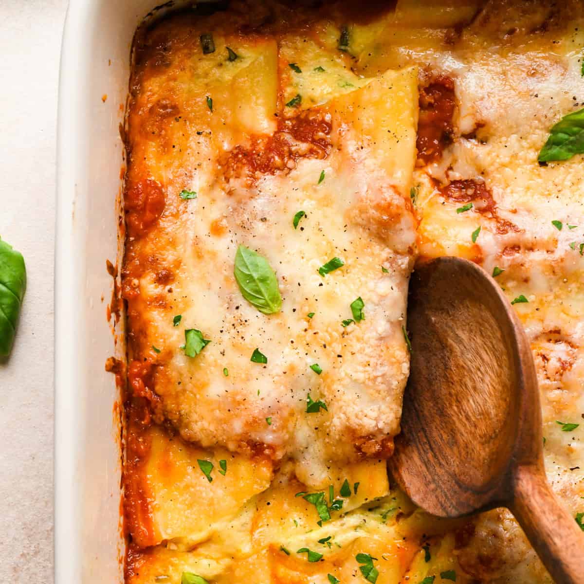 Close-up view of stuffed manicotti in a white casserole dish topped with fresh basil and cracked pepper, being served with a wooden serving spoon.