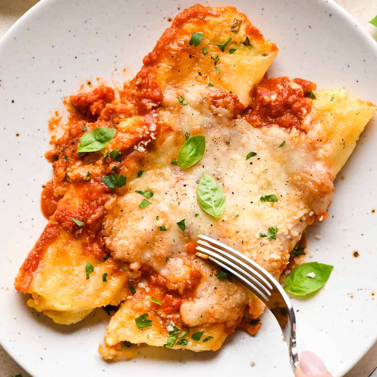 A plate of stuffed manicotti topped with marinara sauce, melted cheese, and fresh basil leaves, served with a fork on a white speckled plate.